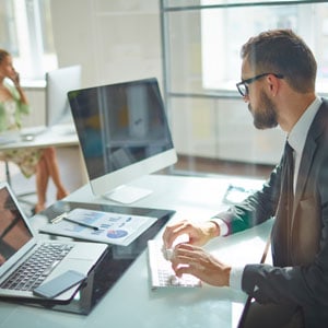 Business professional working at a desk with a computer, laptop, and documents